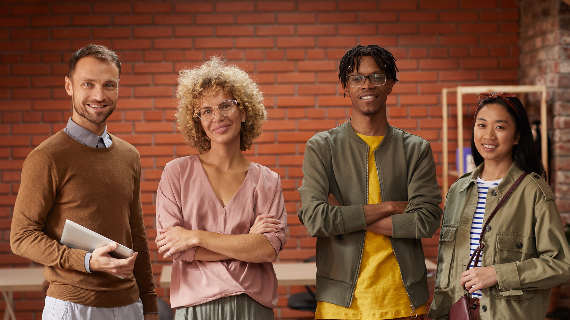 Four team members standing in front of a red brick wall, smiling and looking at the camera in a relaxed office setting.