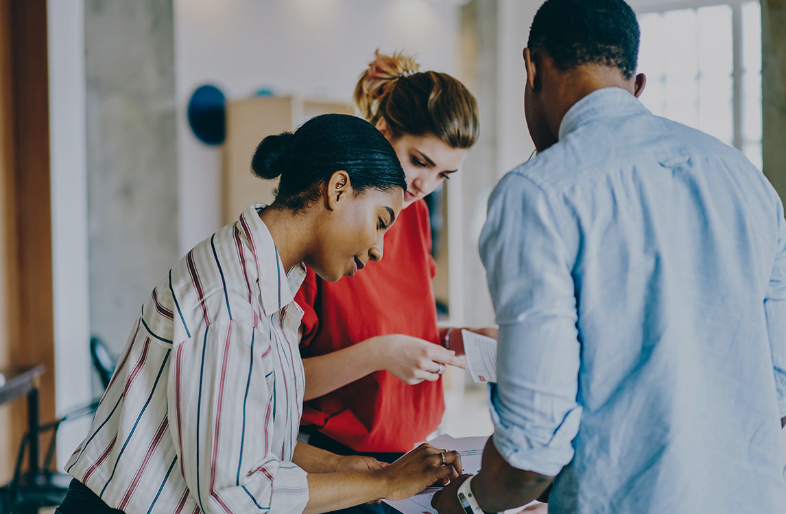 Two professionals working on laptops in a modern café-style workspace with plants and glass walls in the background.