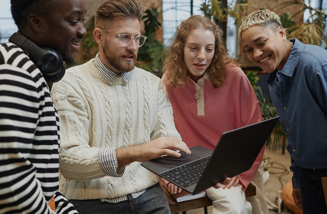 Four colleagues gathered around a laptop in a creative studio, discussing something on screen and smiling together.