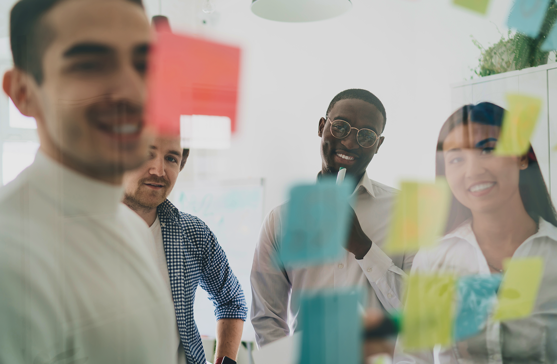 Team of four brainstorming behind a glass wall covered in colourful sticky notes during a creative planning session.