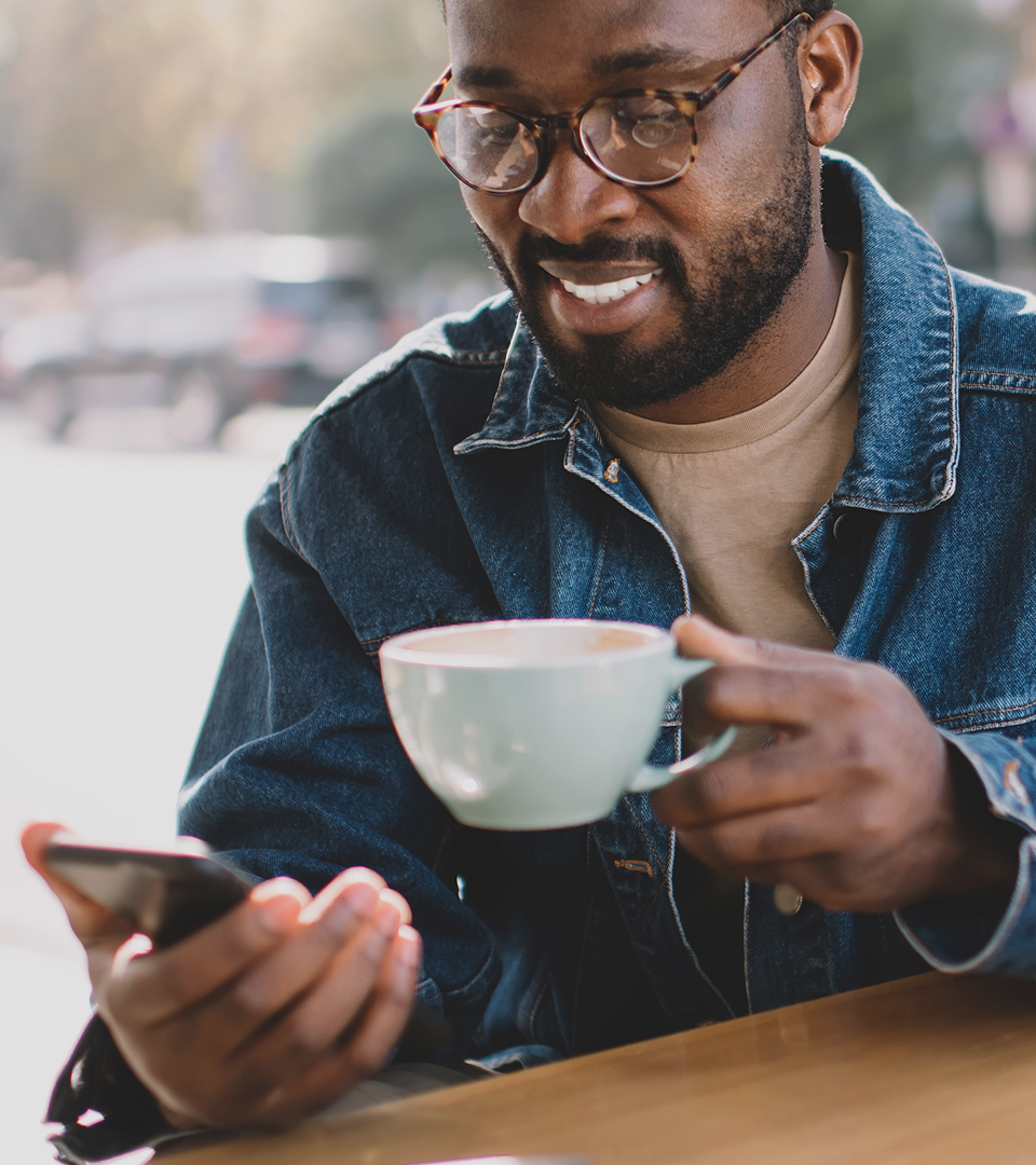 Smiling man sitting at an outdoor café holding a coffee cup and checking his phone, with parked cars in the background.