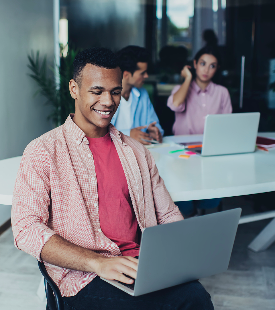 Smiling man working on a laptop in a modern office, with two colleagues having a discussion in the background.