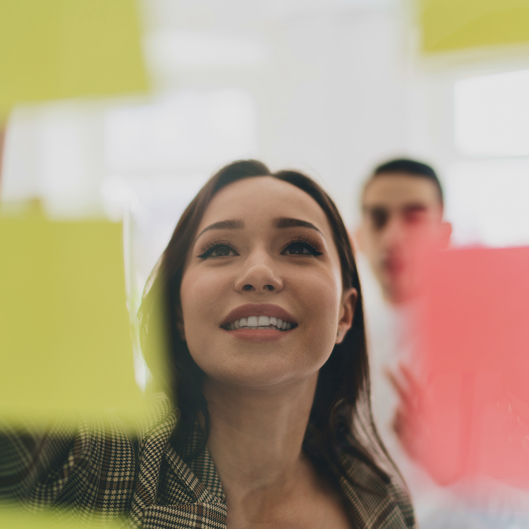 Woman smiling and placing a yellow sticky note on a transparent board while teammates look on during a collaborative workshop.