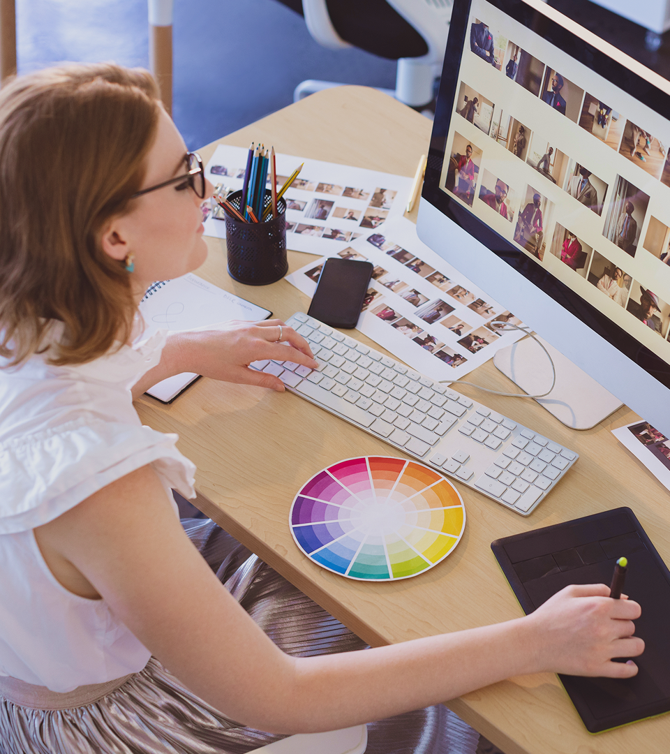 Graphic designer working at a desk with a colour wheel, stylus tablet, and image previews on a large computer screen.
