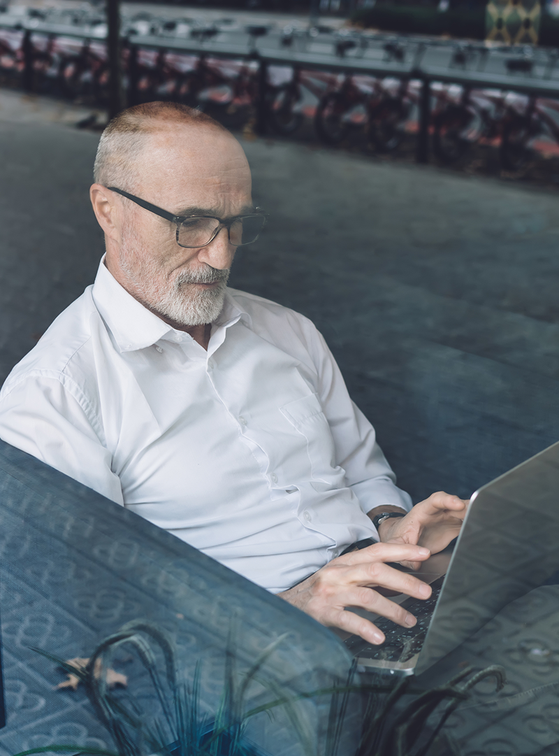 A man working on a laptop at a window seat in an office, seen through the glass from outside.