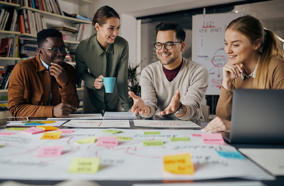 Four teammates smiling and brainstorming around a table covered in charts and sticky notes during a planning session in a studio space.