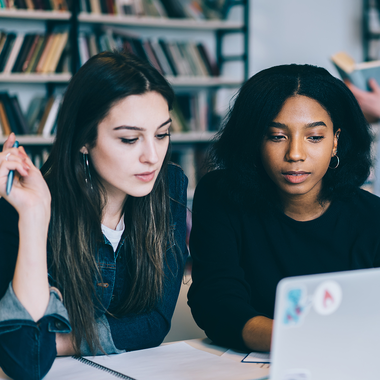 Two women focused on a laptop in an office setting, with notebooks and papers on the table and a man reading in the background.