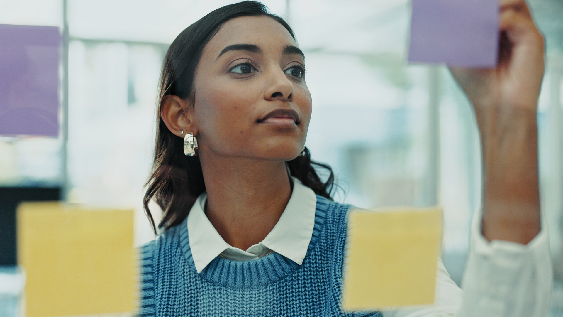 Focused woman placing a purple sticky note on a glass wall during a brainstorming session, surrounded by yellow notes.