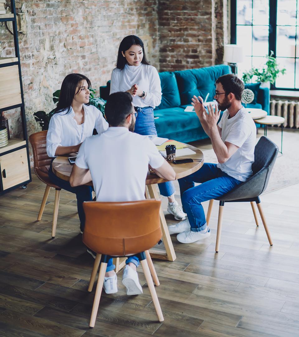 Two professionals discussing work at a desk with a laptop and documents in a bright office with large windows.